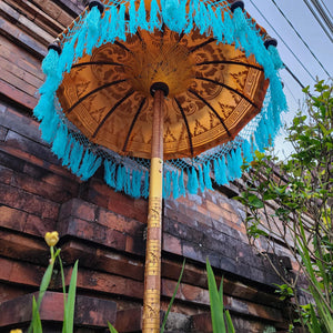 Balinese decorative umbrella with blue fringe and ornate patterns beside a brick wall outdoors