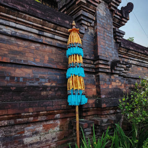 Balinese ceremonial umbrella with golden fabric and blue tassels by a brick temple wall
