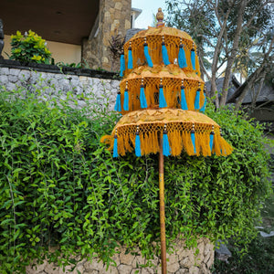 Balinese golden garden parasol with yellow and blue fringe, set against lush greenery outdoors.