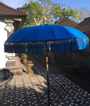 Balinese blue ceremonial umbrella with fringe, outdoors by traditional carved stone decor