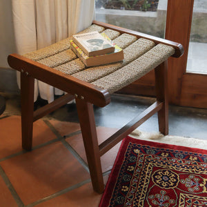 Handcrafted wooden stool with woven seat, book on top, near window and patterned rug