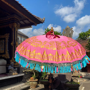Handmade pink Balinese umbrella (Tedung) with intricate gold motifs and blue tassels, set in a tropical environment.