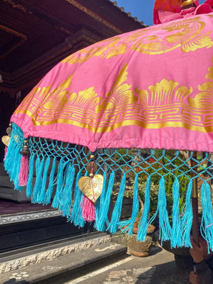 Handmade Balinese umbrella in pink with intricate gold patterns and vibrant blue tassels, perfect for decorating your space.