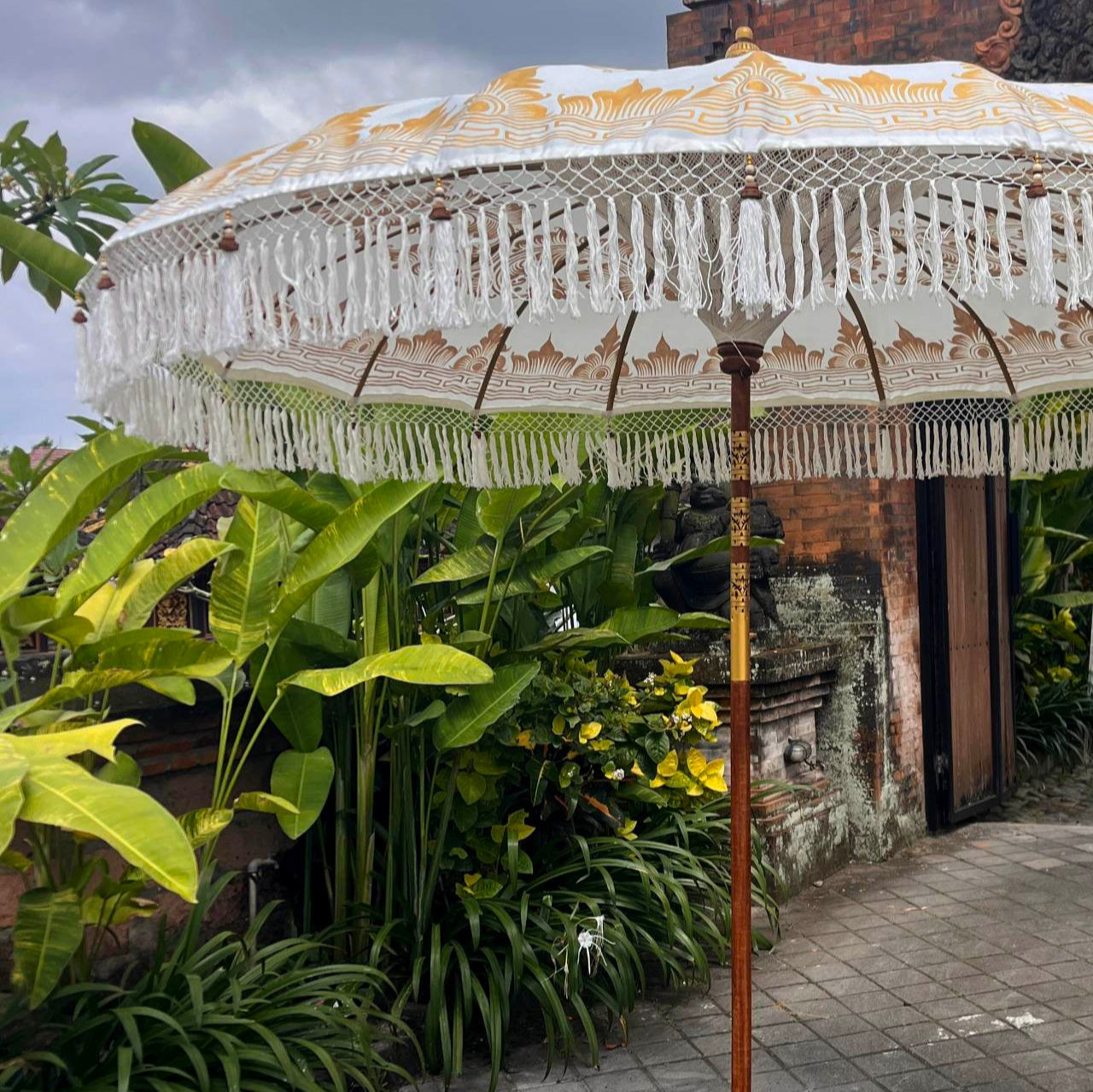 Decorative outdoor umbrella with tassels on a patio with plants and a building in the background