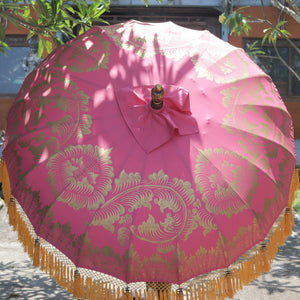 Pink Balinese decorative umbrella with gold patterns and tassels, outdoors in sunlight