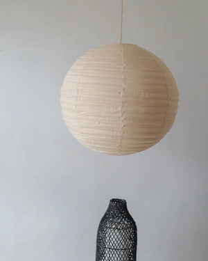 Round beige paper lantern hanging above a black woven decorative vase against a plain wall.