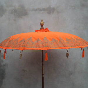 Balinese orange decorative umbrella with tassels and carved wooden pole against gray wall