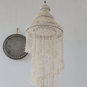 Handmade seashell chandelier with intricate beadwork, hanging on a white wall, next to a decorative round plate with geometric patterns.