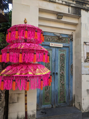 Balinese hand-carved blue door with ornate trim and three pink ceremonial umbrellas.