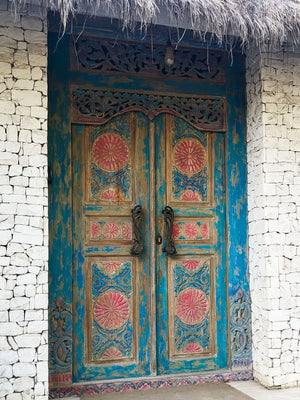 Hand-carved Balinese wooden door with intricate blue and pink floral designs, stone wall