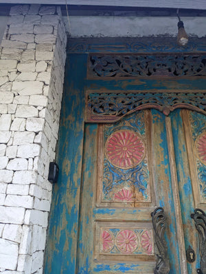 Hand-carved Balinese door with floral motifs and rustic blue, pink, and gold details next to white stone wall.