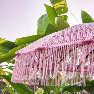 Pink macrame fringe umbrella with woven texture, outdoors among tropical green leaves