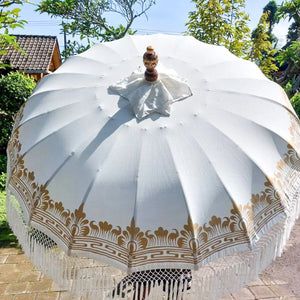 Balinese hand-carved white parasol with gold patterns and fringe, outdoors in garden