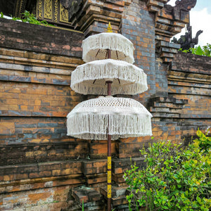 Triple-tier Balinese handmade parasol with white fringe beside a traditional stone wall