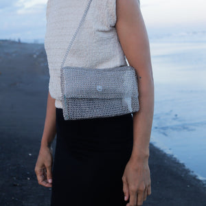 A person standing on a dark beach wearing a silver chain-link crossbody bag with a textured cream sleeveless top and a black skirt.