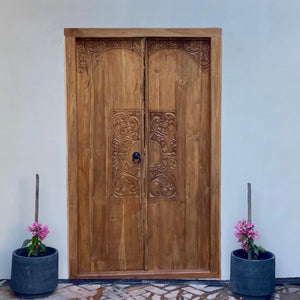 Hand-carved Balinese wooden door with intricate floral motifs and potted pink flowers