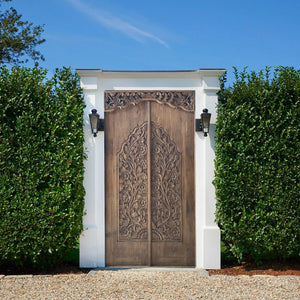 Hand-carved Balinese wooden double door with intricate floral design, set in white frame outdoors.