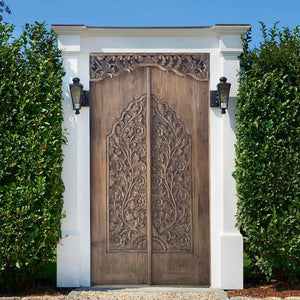 Hand-carved Balinese wooden double door with floral motifs, framed by greenery outdoors