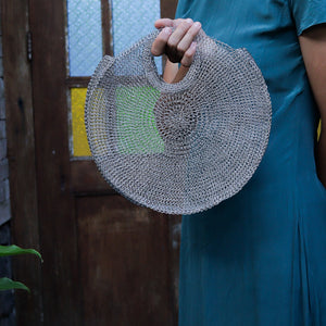 A person in a teal dress holds a large, round, hand-woven straw bag with a circular handle in front of a rustic wooden door.