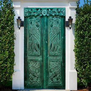 Hand-carved green Balinese door with intricate floral patterns, framed by white pillars and greenery.