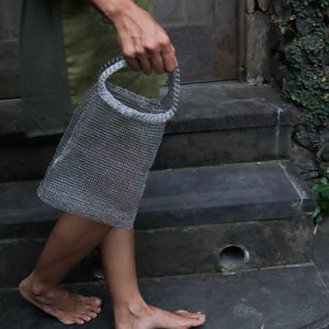 A person holding a small woven mesh handbag with a loop handle, standing on dark stone steps outdoors.