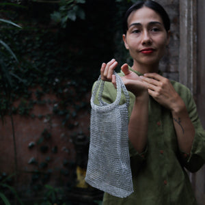 A woman wearing an olive green shirt holds up a gray woven mesh shoulder bag in an outdoor setting with a rustic brick wall and greenery in the background.