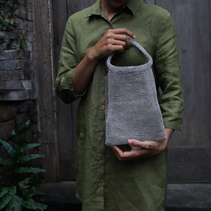 A person wearing an olive green linen shirt holds a small gray knitted shoulder bag with a loop handle, set against a rustic dark wooden door and stone wall background.