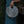 A person in a teal dress holds a round woven straw handbag with a cutout handle, set against a dark green foliage background.