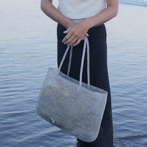 A person standing by the water holds a large woven tote bag in a light gray heathered fabric with matching woven shoulder straps.