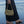 A person standing in shallow ocean water holds a woven shoulder bag with a curved flap and long handles. The person is wearing a long dark skirt, and the beach and water are visible in the background.