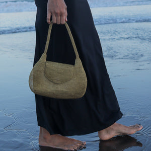 A person standing in shallow ocean water holds a woven shoulder bag with a curved flap and long handles. The person is wearing a long dark skirt, and the beach and water are visible in the background.