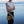 A person standing on a beach at dusk holding a woven straw shoulder bag. The person is wearing a sleeveless textured cream top and black pants, with the ocean and sky in the background.