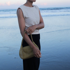 A person standing on a beach at dusk holding a woven straw shoulder bag. The person is wearing a sleeveless textured cream top and black pants, with the ocean and sky in the background.