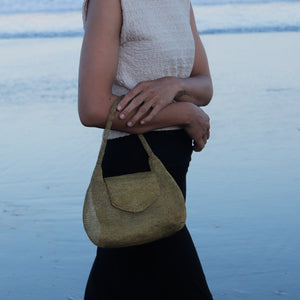 A person standing at a beach holds a woven straw shoulder bag in a light olive green color. The person is wearing a textured cream sleeveless top and a black skirt.