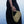 A person wearing a black dress holds a woven straw shoulder bag with a matching woven strap against a soft blue water background.