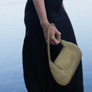 A person wearing a black dress holds a woven straw shoulder bag with a matching woven strap against a soft blue water background.