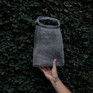 A gray woven mesh handbag with a top handle, held by a hand against a dark green leafy hedge background.