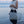 A person standing in shallow ocean water holds a large open-top tote bag made of shimmering silver mesh or woven material with long handles. The background shows the sea and a soft pinkish sky at dusk.