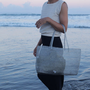 A person standing in shallow ocean water holds a large open-top tote bag made of shimmering silver mesh or woven material with long handles. The background shows the sea and a soft pinkish sky at dusk.