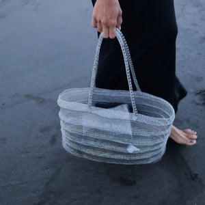Handwoven grey beach bag with striped pattern, carried on dark sand by person in black dress