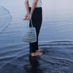 Handwoven seagrass beach bag held by woman standing in ocean waves, eco-friendly coastal accessory