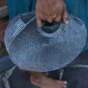 A close-up of a hand holding a large, round woven basket or tray made from silver-colored metal mesh or chainmail-like material, with a person's bare foot visible on a stone surface.