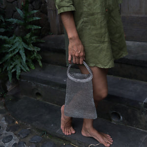 A person wearing an olive green linen dress stands on dark stone steps, holding a small gray chainmail mesh tote bag with a loop handle.