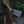 A person outdoors holding a gray woven mesh handbag with a loop handle, wearing an olive green dress on stone steps with green plants in the background.