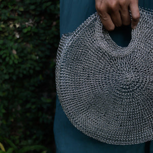 A close-up of a person holding a large, round, silver chainmail-style shoulder bag outdoors, with dark green foliage in the background.