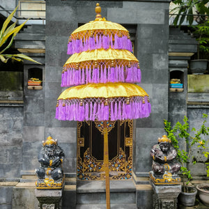 Decorative umbrella with purple fringe and gold accents in front of a stone wall with statues and plants.