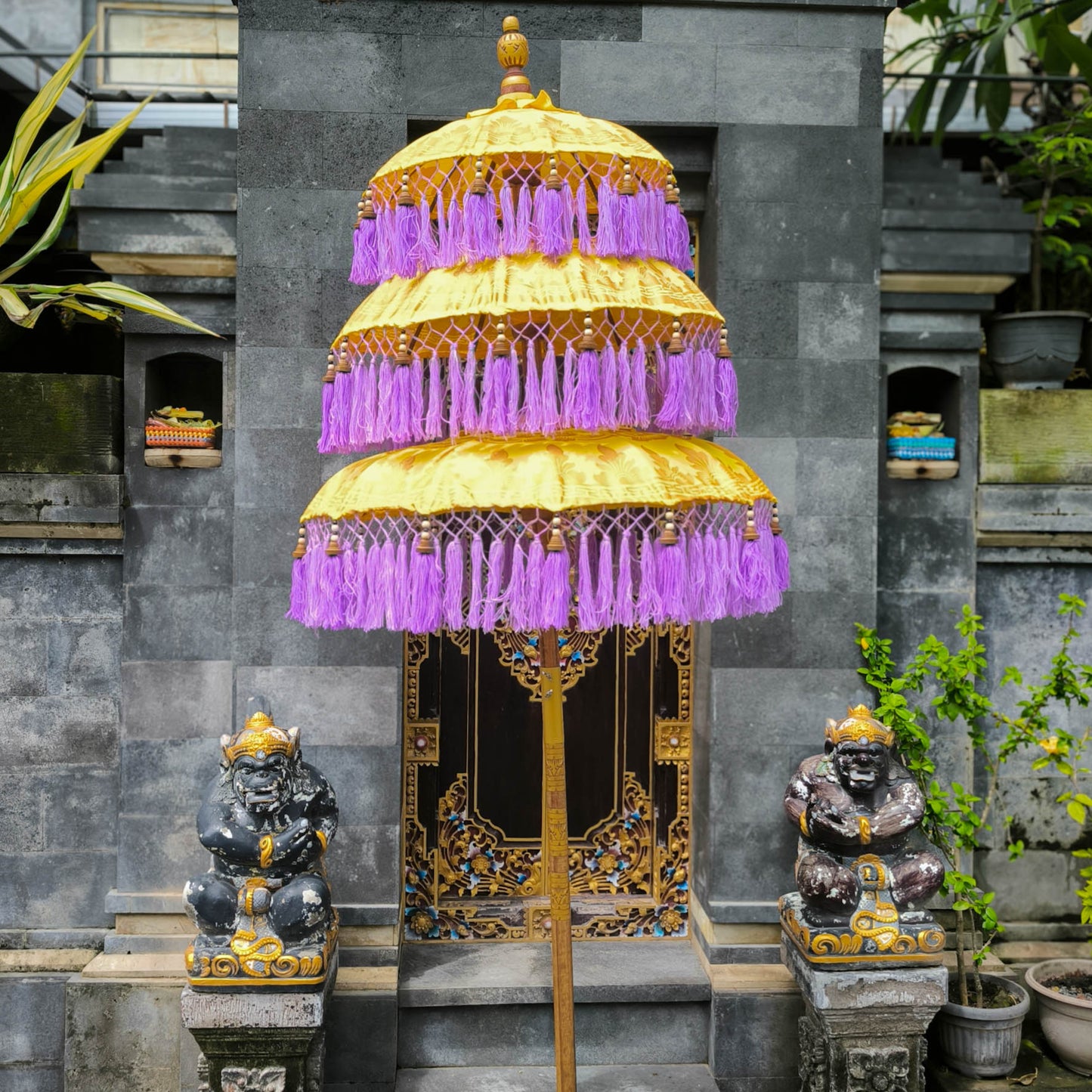Decorative umbrella with purple fringe and gold accents in front of a stone wall with statues and plants.