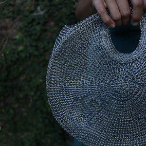 A close-up of a person holding a large, round woven bag made from silver-toned chainmail or mesh links, with a dark green foliage background.