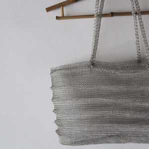 A silver metallic woven mesh tote bag hanging from a wooden rod against a plain white wall.