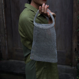 A person wearing an olive green shirt holds up a gray woven mesh shoulder bag with a single top handle, set against a dark rustic wooden door background.